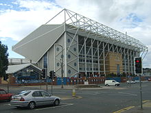 Outside a large stadium with a cantilevered stand at the right-hand end and some lower buildings on the left.