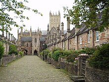 Street scene. Terraces of old red brick houses on either side of the road. At the far end is a building with arches and small tower. Beyond can be seen the cathedral tower.