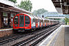 Passenger train of 1992 stock. The train's cars are white, with red sliding doors, with the bottom half-metre of the sides blue. The train is at the platform of an open-air station.