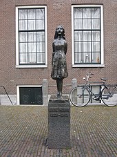 A bronze statue of a smiling Anne Frank, wearing a short dress and standing with her arms behind her back, sits upon a stone plinth with a plaque reading "Anne Frank 1929–1945". The statue is in a small square, and behind it is a brick building with two large windows, and a bicycle. The statue stands between the two windows.