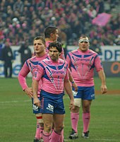 Au premier plan quatre joueurs sont statiques lors d'un temps mort, vêtus de rose et de bleu. Au fond, on devine les spectateurs parisiens avec les mêmes couleurs en tribune.