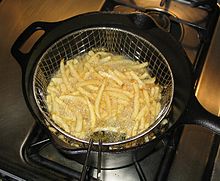 Gros plan sur une casserole en fonte posée sur un fourneau à gaz. Dans le récipient rempli d’huile chaude, un panier métallique grillagé à manche contient les frites qui dorent ; entre le panier et la paroi de la casserole, on voit roussir des petits morceaux de frite qui se sont échappés des mailles du panier.