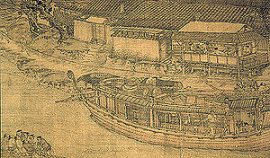 A close up view of a large trading barge crewed by multiple people. The barge has wooden walls surrounding it on all sides, and a thin line of tiled roof capping the walls, but not covering the interior of the ship. There are several windows built into the wall of the vessel.