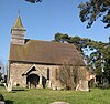 A stone church with a tiled roof seen from the south. On the left gable is a shingled bellcote, and a wooden porch protrudes from the church