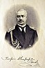 Black and white head and shoulders photograph portrait of a man in a black jacket and tie, with epaulettes and braid