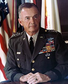 White-haired, clean-shaven man in dark-green ceremonial military uniform and tie. He has a lot of insignia on the left side of his chest and stars to indicate that he is a general.