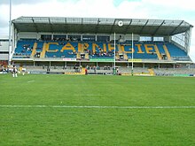 Inside a stadium with grass in the foreground and rugby posts and a large three-storey roofed stand with clock at the far end. At ground-level there are steps and railings for standing patrons, and in front are a number of advertisements for the Leeds Building Society, PowerGen, Studio Jeff Banks, etc. The upper part of the stand has about fifteen rows of seats, most of them coloured blue but the others collectively spelling out the word "Carnegie". There are a small number of patrons in both sections and a few more on the top level in front of some windows at the rear. To the left of the stand is part of another three-storey building. In front of this, at the far left-hand end of the pitch, are a few drum-majorettes with pompoms.