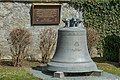 Cloche posée sur un piédestal en bois sur un terrain de gazon avec un mur blanc en arrière-plan avec une plaque commémorative en métal.