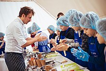 Yevhen Klopotenko speaking to a group of children wearing hairnets and aprons at a table with cooking equipment