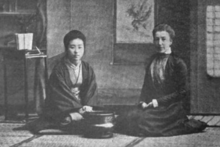 Two women are seated on the floor in a Japanese-style room. The woman on the left is Japanese, wearing a shawl; the woman on the right is white, wearing western dress typical of the late nineteenth century.