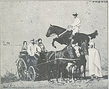 Photo en noir et blanc d'un cavalier monté sur un cheval noir sautant au-dessus d'un attelage de trois chevaux avec quatre personnes à bord