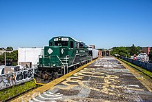 Dunkelgrüne Diesellok Nummer 300 mit weißen Beschriftungen NEW YORK AND ATLANTIC RAILWAY. Verfallener Mittelbahnsteig erhöht auf Viadukt/Bahndamm, funktionsloses Dach im Hintergrund, links und rechts leicht grün bewachsene Gleise und jenseits Gebäude der Stadt