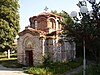 Side view of a small Byzantine-style medieval church constructed out of bricks and mortar. The church's entrance is visible. A street lamp and greenery in the surrounding area.