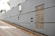 Seitenmauer einer Tiefgaragenabfahrt, die Mauer ist verputzt und grau gestrichen, ein Rechteck ist ausgespart und erlaubt den Blick auf die brutalistische Betonmauer und ein sehr schemenhaft erhaltenes Graffiti. Auf dem Boden liegt Herbstlaub