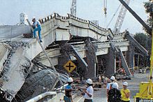 A collapsed elevated freeway with the upper deck resting on the lower deck and several pillars destroyed. Several people with hard hats are seen investigating the scene.