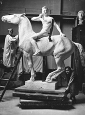 The artist places her left hand on the neck of the model of a horse whose head is above hers