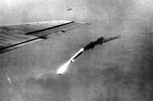 Black and white photo of flaming wreckage falling towards the ground. The wing of a plane is visible at the left-hand side of the photo.