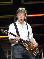 Colour photograph of McCartney, in his sixties, playing a Höfner 500/1 electric bass. He wears a black buttoned-up suit jacket with black pants.