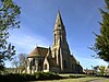 A stone church seen from the northeast with a broad tower and spire, a chancel with an apse and, between them a small vestry