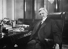 A man sitting at a desk looking directly at the photographer; one hand is holding a pen to a document on his desk while the other is hanging over the arm of his high backed chair, his legged stretched forwards, he is wearing a suit, necktie, and glasses, and has a serious expression.