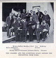 A group of 16 men, standing or sitting, on the deck of a ship with a small lifeboat visible, left background. The group's pose is casual and the men are variously attired, many in suits with casual hats of different sorts.