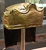 A gold-tinted metal helmet seen from the front right, with three peaks on the brow band, bordered with snakes, and three figures embossed and engraved upon it