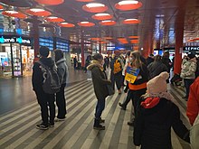 People with yellow and blue signs reading "refugee help" in a station with shops