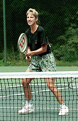 A blonde-haired female tennis player with multi-coloured shorts and a black shirt, with the tennis racket out in front of her