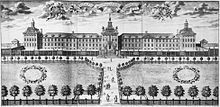 A rather massive facade of a very wide building with two wings projecting from a central structure. It is enclosed by low walls in front of which there are decorative gardens.