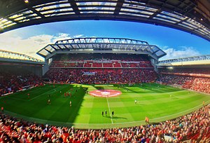 Three football stands with red seats filling up with spectators and surrounding a pitch.