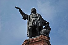 Sculpture of a man in European clothes. He holds one hand to the sky and, with the other, takes a veil off of the globe.