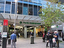 An entrance to the underground station, with escalators and stairs going down. The entrance building has another building built on top and around it. A large sign is on top of the entrance, which reads "Perth Underground". There is a crowd of people in and around the entrance.
