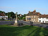 Stone cross surrounded by railings on grass area in front of roads and houses.