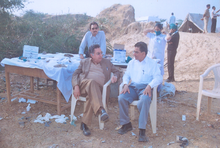 Two men seated outdoors, with other men standing