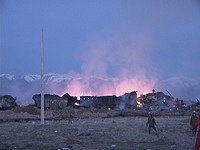 In twilight against snow capped mountains, two firefighters walk through a field flanked by burning rubble of the village