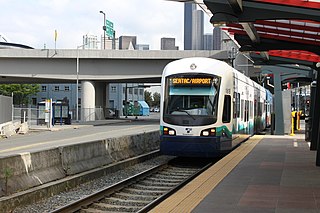 Modrobílá tramvaj přijíždějící do stanice. Stanice má dlážděnou podlahu, kterou chrání prosklená černočervená střecha. Po levé straně od kolejnice vede silniční komunikace, nad níž stojí most. V pozadí snímku se tyčí několik mrakodrapů.