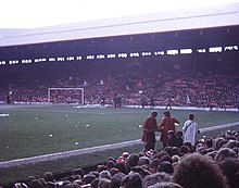 Photograph of a stand full of people some of whom are waving flags. In front of the stand is a field and a goal.