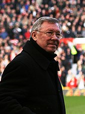 The torso and head of a grey-haired white man. He is wearing spectacles and a black coat.