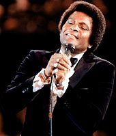 A dark-skinned man in a tuxedo singing into a microphone