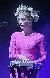 A picture of Lorde on stage wearing a pink dress and flower crown as plays a MIDI controller.