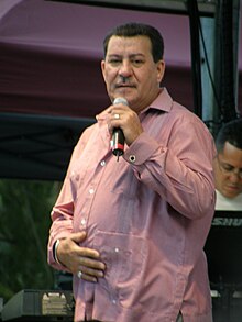 A man wearing a pink button-shirt is holding a microphone on his left hand.