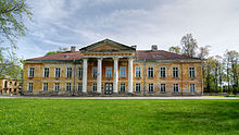 Frontale Farbfotografie eines heruntergekommenen Gebäudes mit einem Säuleneingang und einem Giebelfeld. Die Fassade hat einen schmutzigen Braunton und die Steine sind im unteren Bereich zu sehen. Seitlich sind Bäume und im Vordergrund ist eine Wiese.