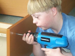 A Caucasian adolescent male with Down syndrome dressed in a blue t-shirt is holding a blue and black electric drill with both hands. He is using it to assemble a wooden, flat-pack style bookcase.