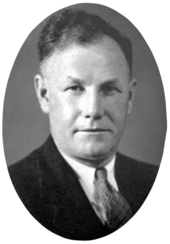 A head and shoulders, black and white photographic portrait of a white man with receding dark hair