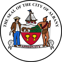 A black circle with white fill is shown with the coat of arms at center. Above the coat of arms, following an arced path, are the letters "The Seal of the City of Albany"