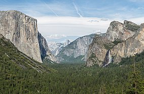 Mountain valley with sheer cliff on its left side, and a waterfall cascading into its right, with a clear blue sky above and many green trees below