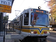 A white and yellow light rail train at a station platform with overhead lines visible above.