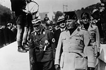 A group of men in formal and paramilitary clothing walk past a row of boys in uniforms