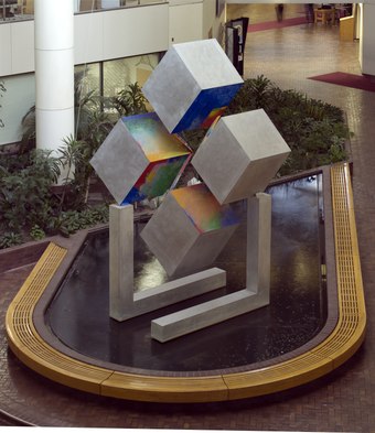 A photograph of a seventeen-foot tall aluminum sculpture of flattened cubelike faces standing on two angular legs. The interior faces of each cube have been painted in rainbow colors.