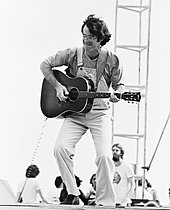 A man with dark hair and a mustache, wearing dungarees, playing a guitar on a stage